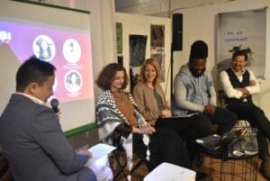 This photo features our side event at the International Journalism festival. You see five individuals sitting in chairs in front of projector screen. They are engaged in conversation with our event moderator.