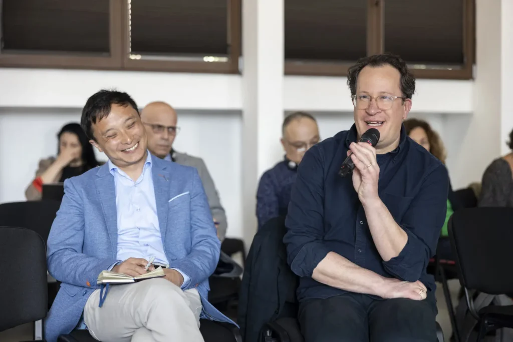 Lei Ma and Boris van Westering attend Assise du Journalisme. Lei sits beside Boris, who is using a microphone to speak.